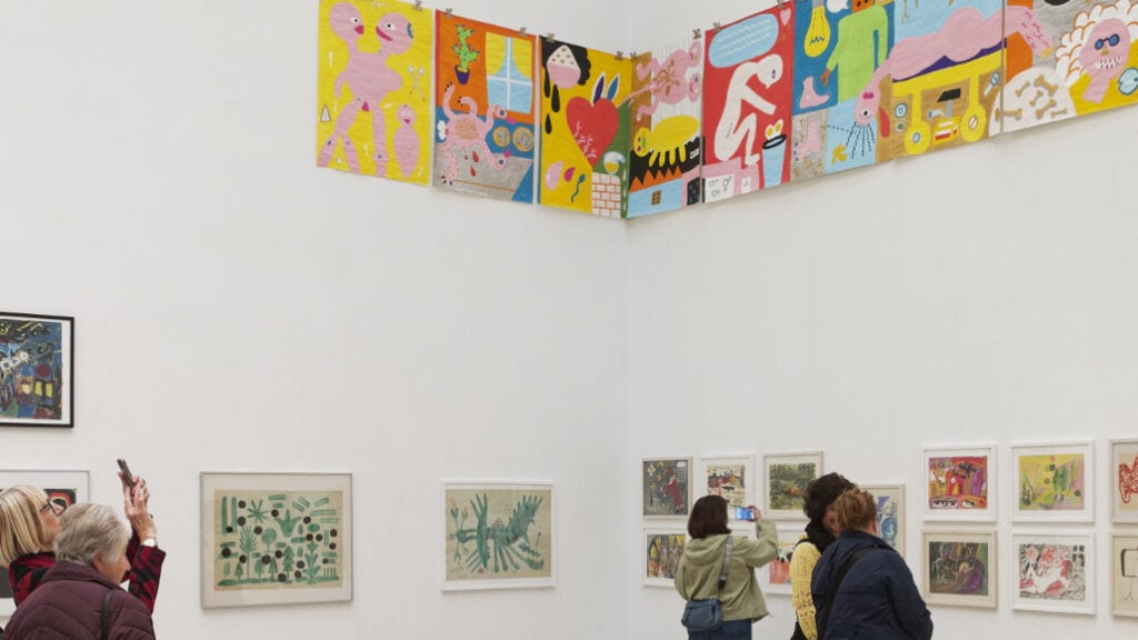 Ausstellungsansicht mit farbenfrohen Kunstwerken von Holger John an weißen Galeriewänden, während Besucherinnen die Werke betrachten und fotografieren.
