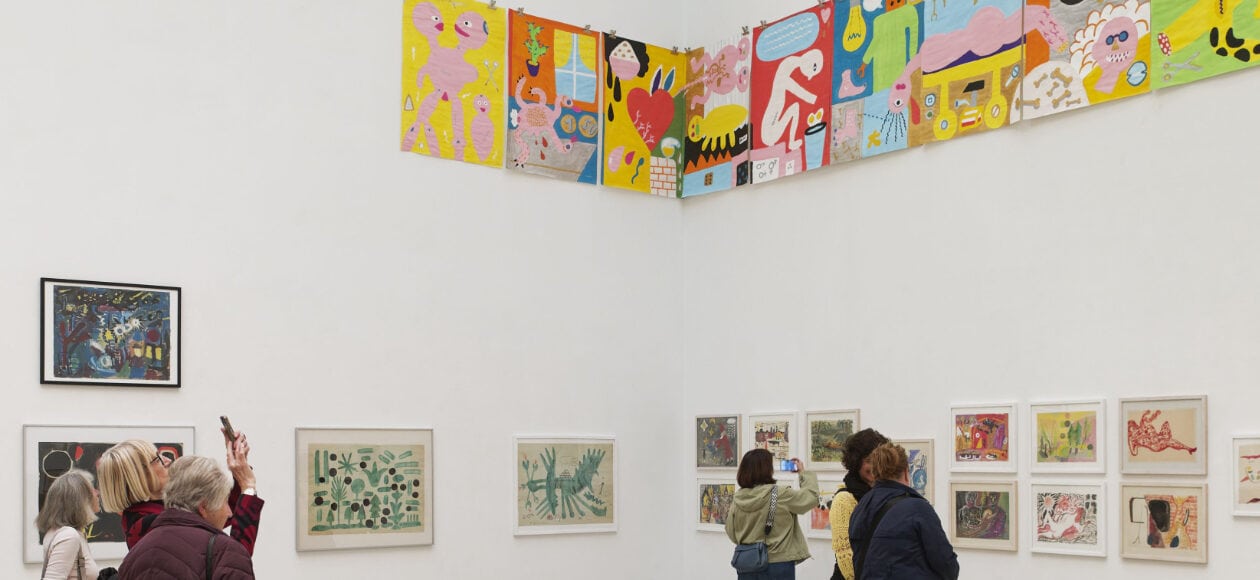 Ausstellungsansicht mit farbenfrohen Kunstwerken von Holger John an weißen Galeriewänden, während Besucherinnen die Werke betrachten und fotografieren.