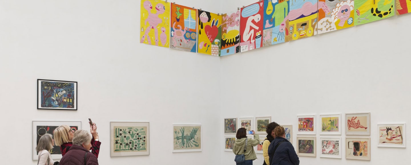Ausstellungsansicht mit farbenfrohen Kunstwerken von Holger John an weißen Galeriewänden, während Besucherinnen die Werke betrachten und fotografieren.