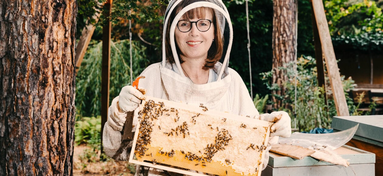 Imkerin im weißen Schutzanzug hält Honigwabe voller Bienen in grüner Gartenlandschaft.