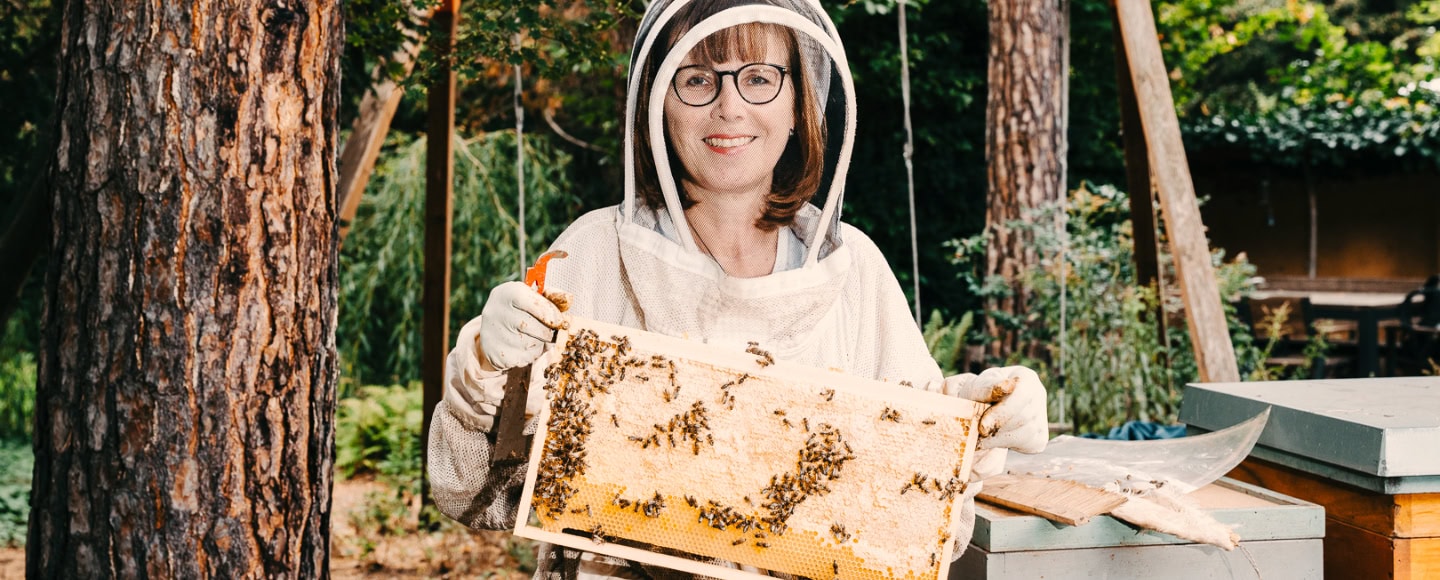 Imkerin im weißen Schutzanzug hält Honigwabe voller Bienen in grüner Gartenlandschaft.