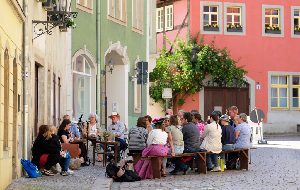 Besucherinnen und Besucher des Literaturfestes Meißen sitzen in einer Seitenstraße auf Bänken und lauschen einer Lesung.