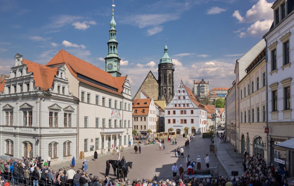 Belebter Marktplatz der Stadt Pirna mit historischen Giebelhäusern, Kirchturm und Zuschauermenge unter blauem Himmel.  