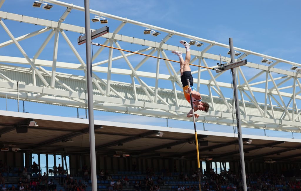 Stabhochspringer in orange-weißer Kleidung überschreitet die Latte im modernen Leichtathletikstadion Goldenes Oval Dresden.