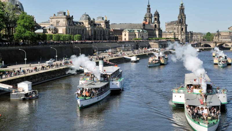 Mehrere weiße Raddampfer der sächsischen Dampfschiffahrt mit schwarzem Rauch fahren auf der Elbe vor der Dresdner Altstadt und steinernen Uferpromenade.