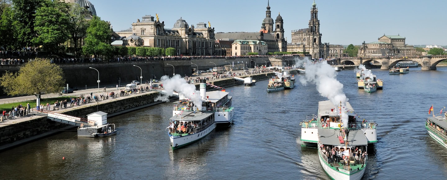 Mehrere weiße Raddampfer der sächsischen Dampfschiffahrt mit schwarzem Rauch fahren auf der Elbe vor der Dresdner Altstadt und steinernen Uferpromenade.