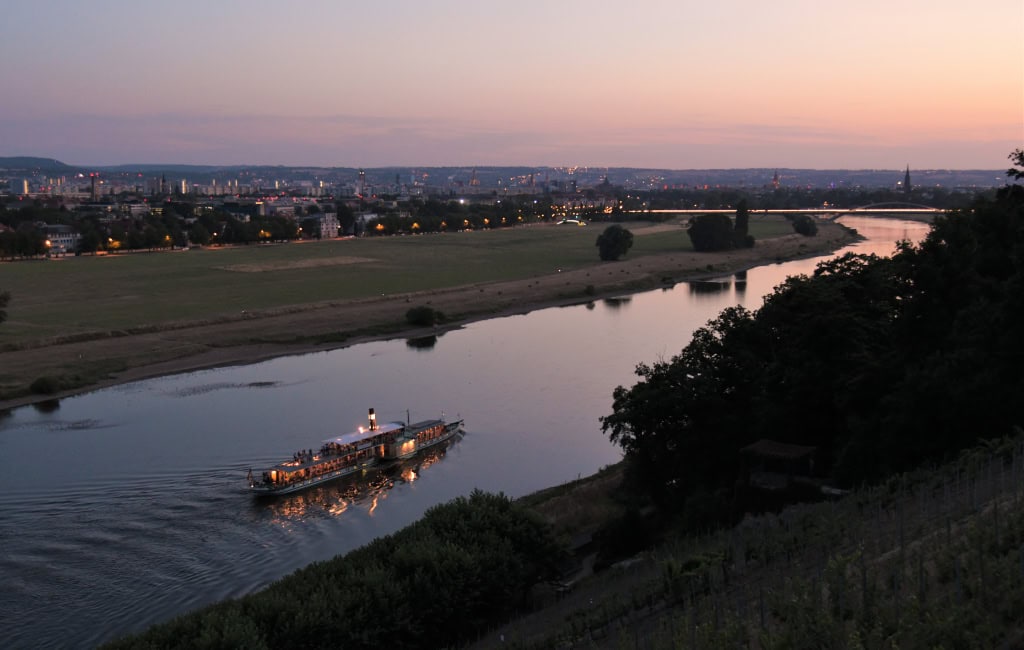 Beleuchteter Raddampfer der Weißen Flotte Dresden fährt bei rosafarbenem Abendhimmel auf der Elbe an dunklen Hängen und Dresdner Wiesen vorbei.