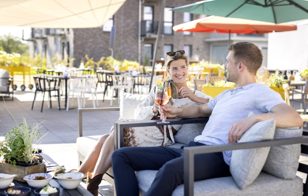 Feierabend Dresden: Zwei Gäste mit Aperitif auf grauem Loungesofa unter bunten Sonnenschirmen auf einer Terrasse