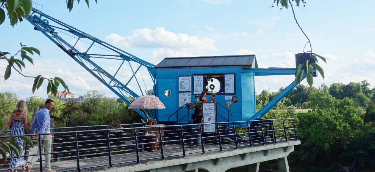 Blauer Hafenkran mit Caféfenster auf Plattform, umgeben von Geländer, Bäumen und blauem Himmel.“