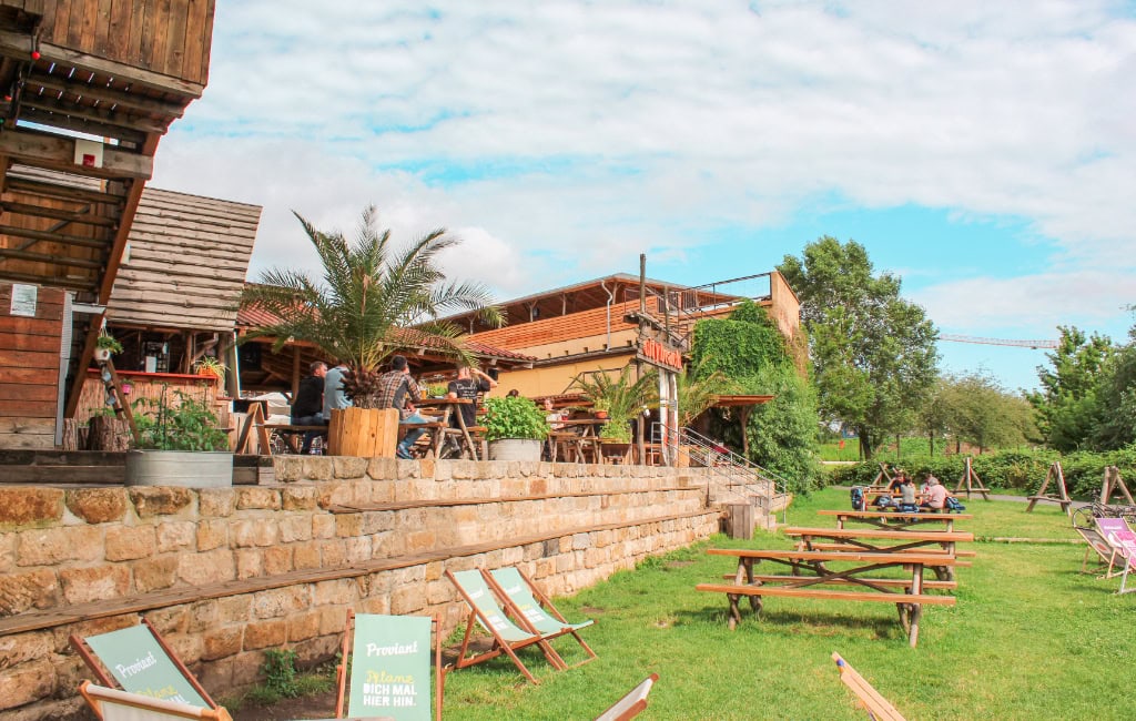 Feierabend in Dresden: Holzterrasse mit Palmen, Sitzplätzen und Wiese vor rustikalem Gastrogebäude am Citybeach.