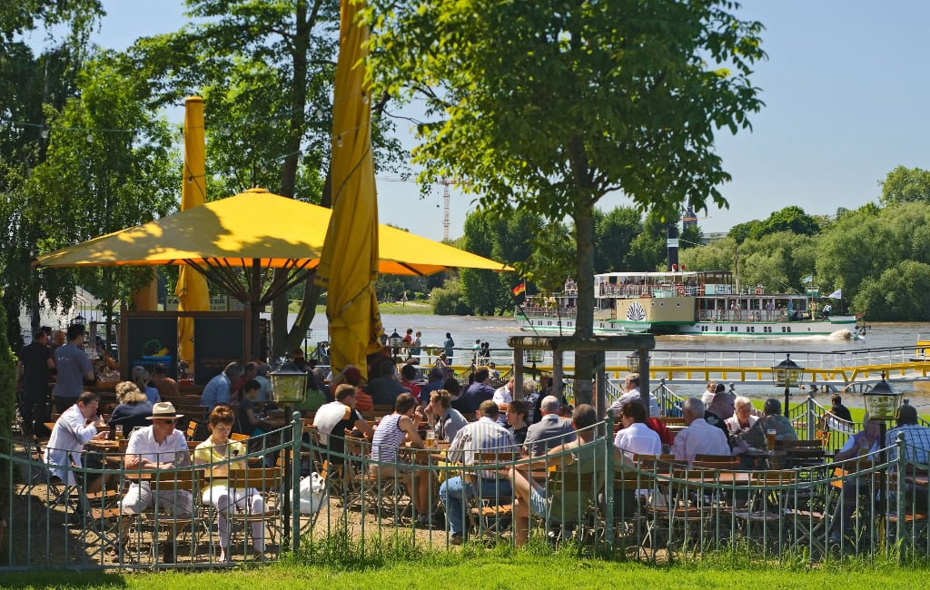 Feierabend Dresden: Biergarten am Elbufer mit gelben Sonnenschirmen, vielen Tischen und Raddampfer im Fluss.