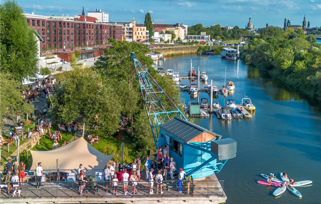 Blick über den Neustädter Hafen mit blauem Kran, Booten am Steg und Flusslauf zur Altstadt.