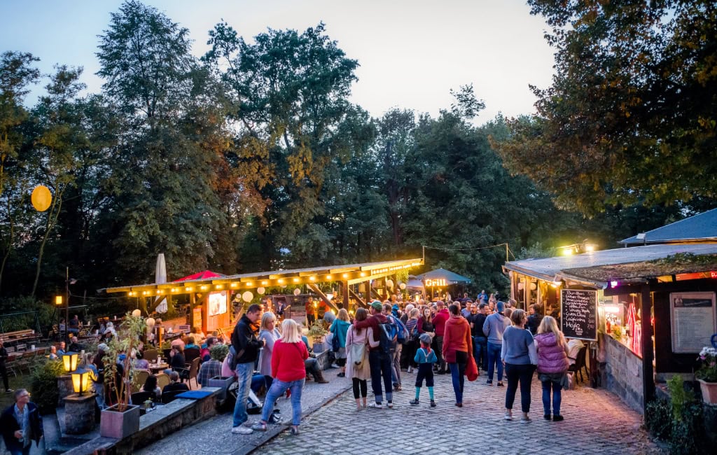 Beleuchteter Biergarten Saloppe mit vielen Gästen, Marktständen und Bäumen in der Abenddämmerung