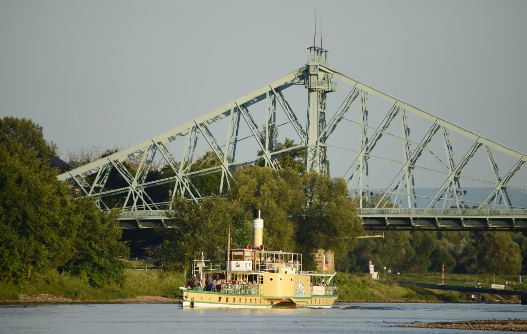 Cremefarbener Raddampfer der sächsischen Dampfschiffahrt  fährt auf der Elbe vor dem hellblauen Stahlfachwerk der Brücke Blaues Wunder.