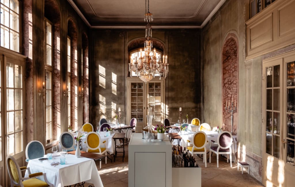 Restaurants in Dresden: Eleganter Speisesaal mit Kronleuchter, hohen Fenstern und bunt gepolsterten Stühlen.