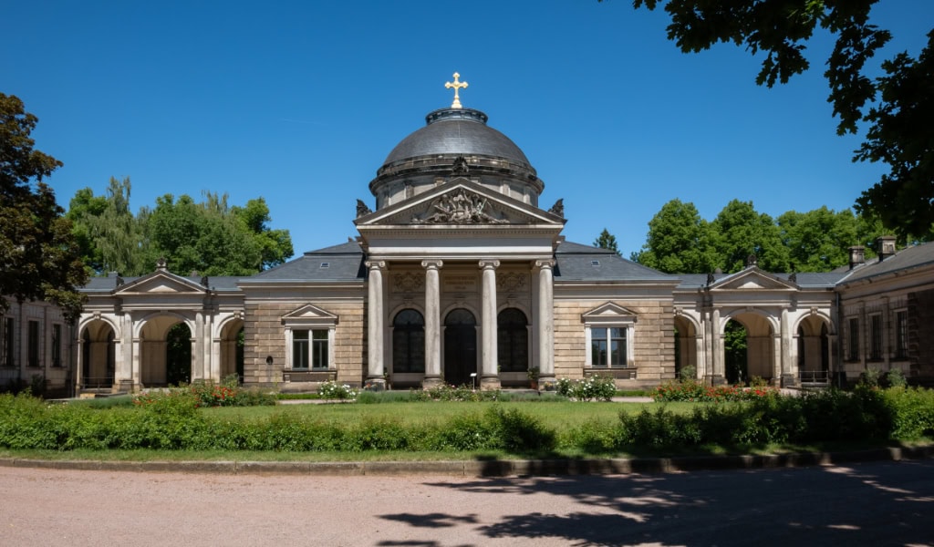 Symmetrische Trauerhalle mit zentraler Kuppel, Kreuz, Säulenportikus und langen Seitenflügeln vor blauem Sommerhimmel