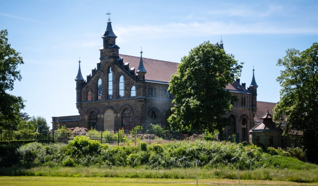 Historisches Backsteingebäude mit vielen Spitztürmen und hohen Fenstern, umgeben von Bäumen und grüner Wiese  