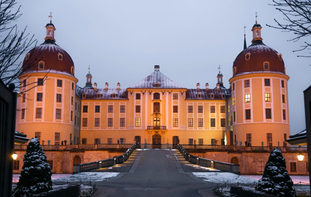Das barocke Schloss Moritzburg bei Dresden erstrahlt in der Abenddämmerung. Schnee bedeckt die Dächer und die Schlosszufahrt.