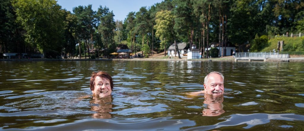 Ein älteres Paar badet im Natursee der Badestelle Weixdorf, im Hintergrund sind Ufer, Bäume und Gebäude zu sehen.