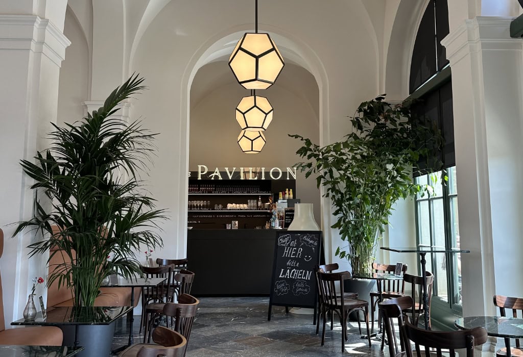 nnenbereich des Café The Pavilion in Dresden mit hohen Rundbögen, großen Grünpflanzen, Sitzgruppen und geometrischen Hängeleuchten.
