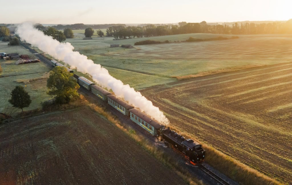 Dresden für Kinder: Die historische Lößnitzgrundbahn dampft im goldenen Morgenlicht durch die weite Landschaft des Elblands bei Dresden.