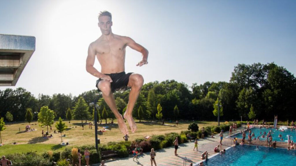 Ein Mann springt vom Sprungturm ins Schwimmbecken des Freibads Wostra, im Hintergrund entspannen Badegäste auf der Liegewiese.