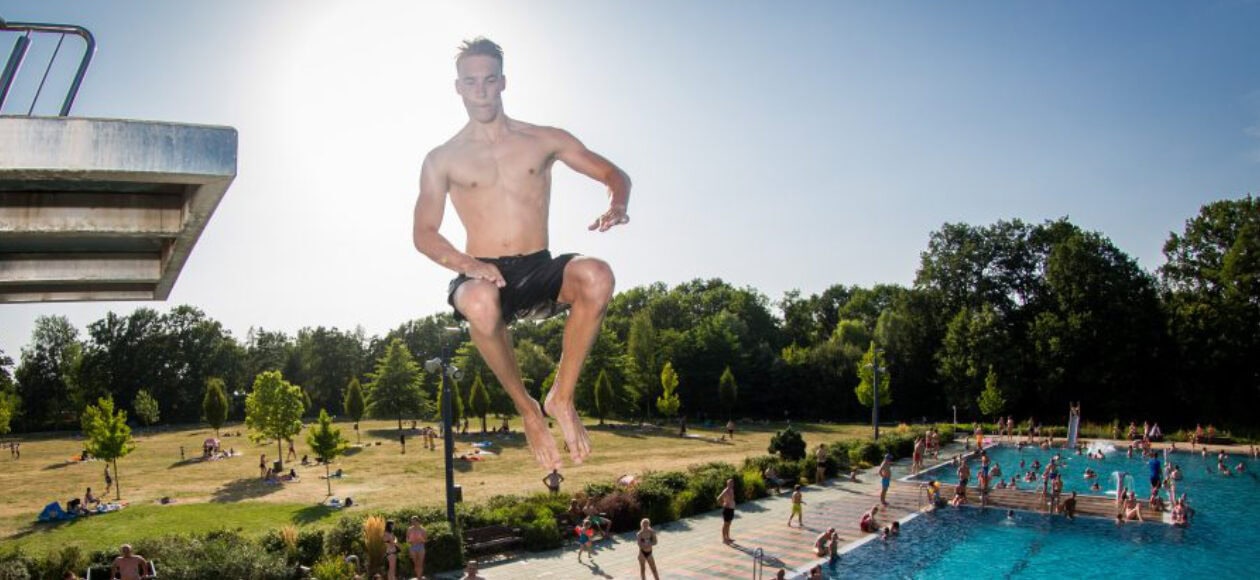 Ein Mann springt vom Sprungturm ins Schwimmbecken des Freibads Wostra, im Hintergrund entspannen Badegäste auf der Liegewiese.