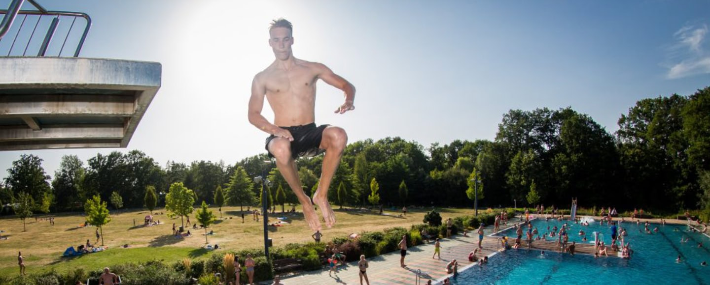 Ein Mann springt vom Sprungturm ins Schwimmbecken des Freibads Wostra, im Hintergrund entspannen Badegäste auf der Liegewiese.