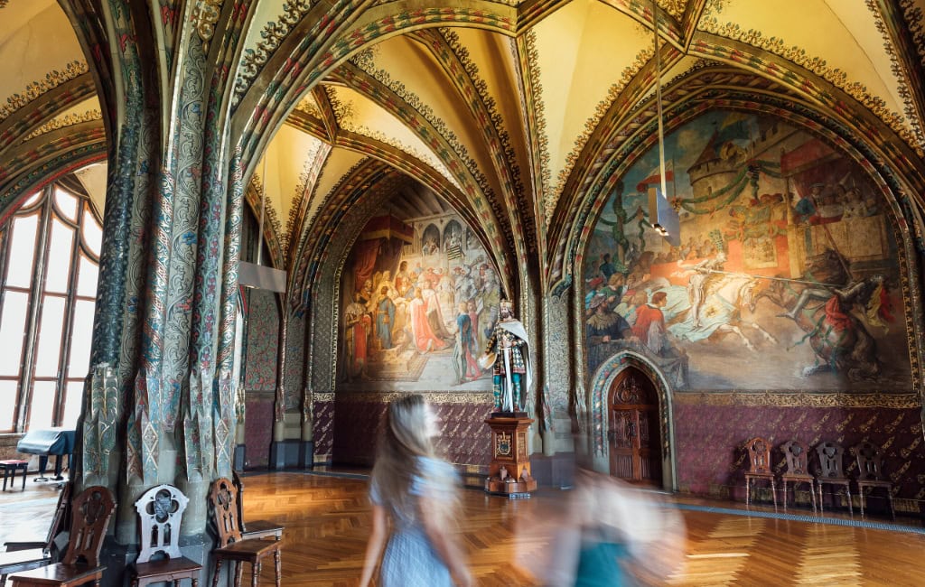 Prächtiger Saal der Albrechtsburg Meißen mit kunstvoll verzierten Gewölbedecken, farbenfrohen Wandgemälden und Besucherinnen im Vordergrund.