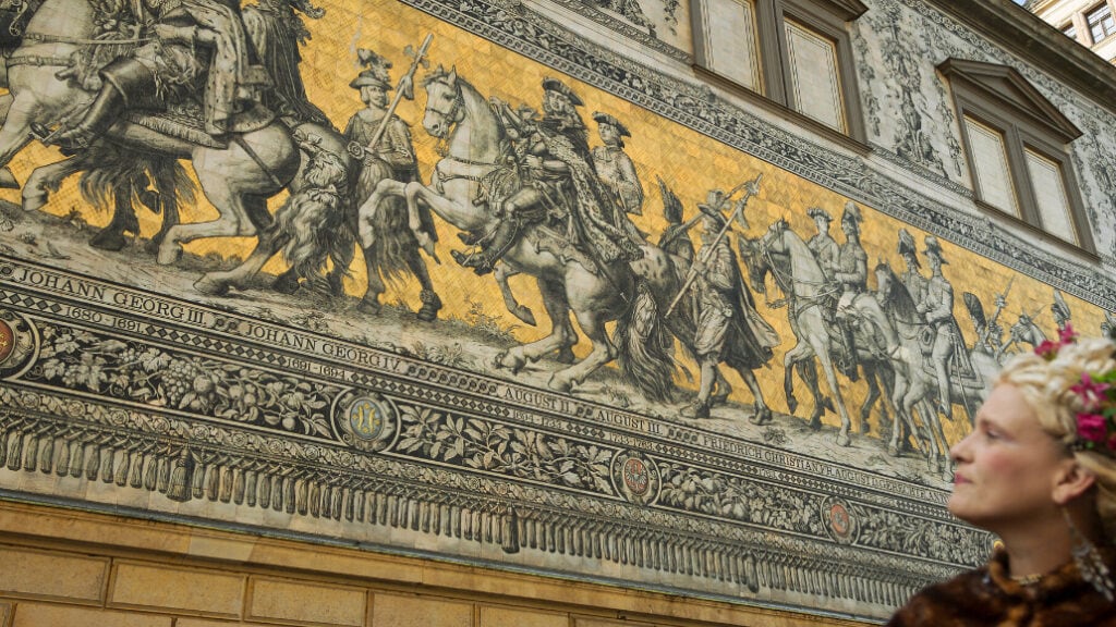Historischer Fürstenzug in Dresden mit goldfarbenem Wandbild und einer Frau in barockem Kostüm im Vordergrund.