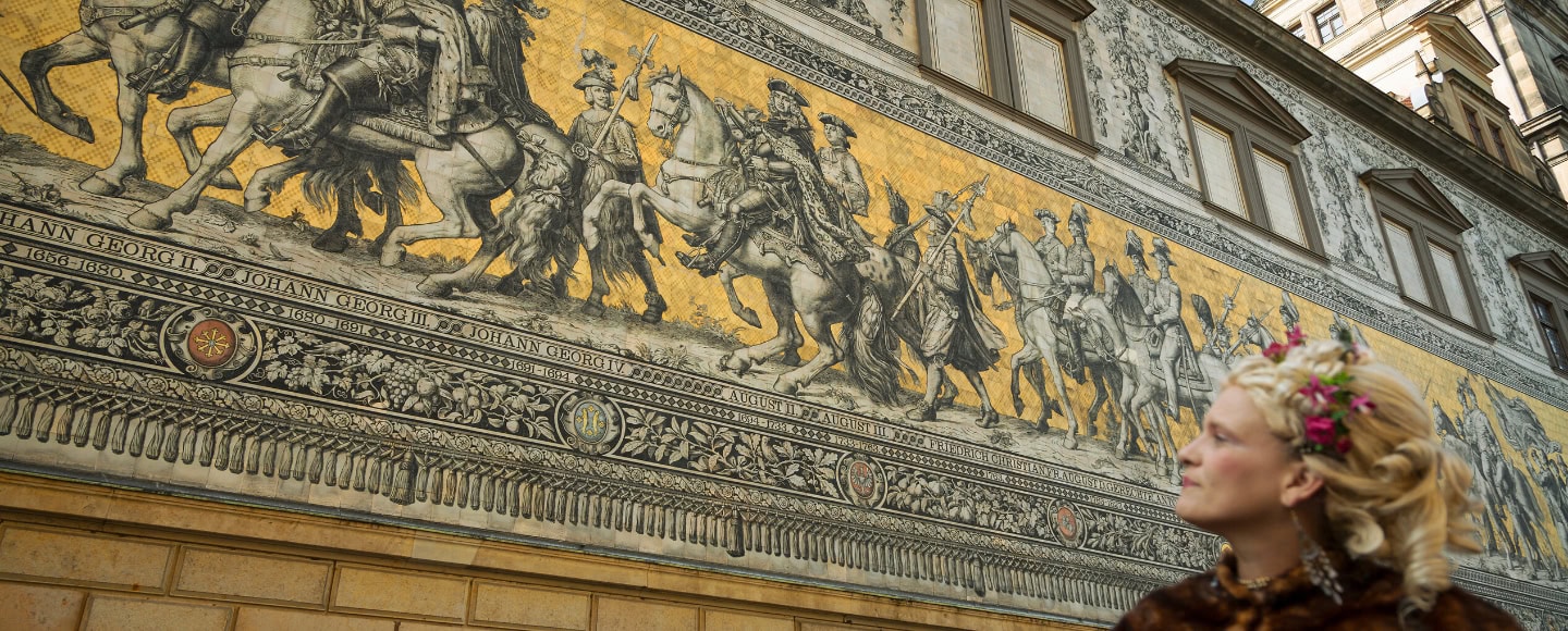 Historischer Fürstenzug in Dresden mit goldfarbenem Wandbild und einer Frau in barockem Kostüm im Vordergrund.