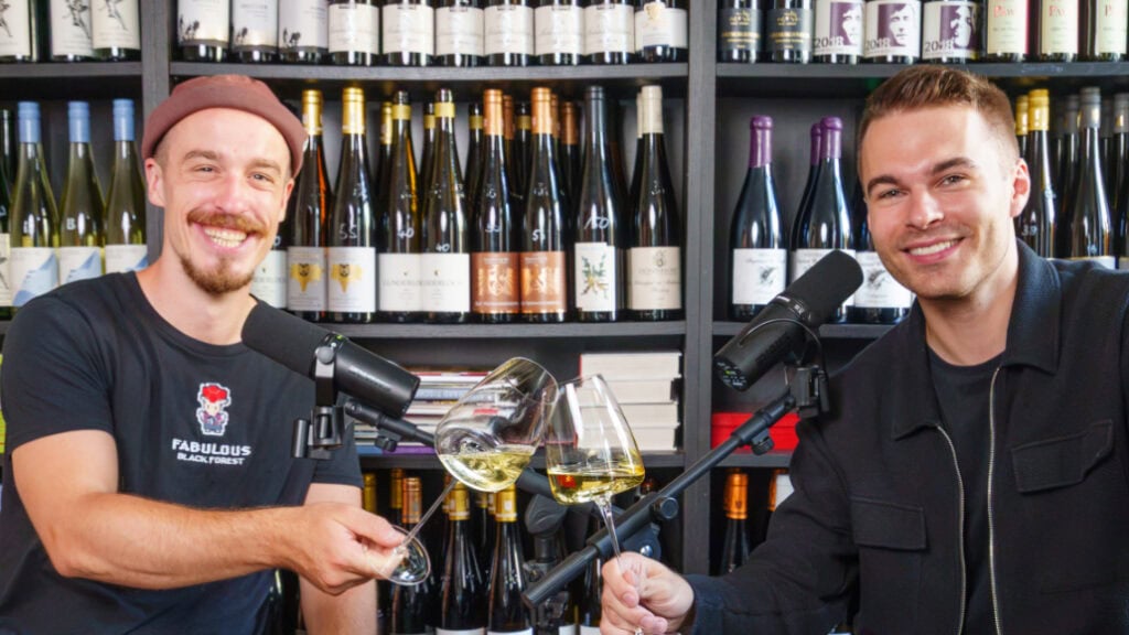 Zwei Männer sitzen vor einem Weinregal und stoßen mit einem Glas Weißwein an.