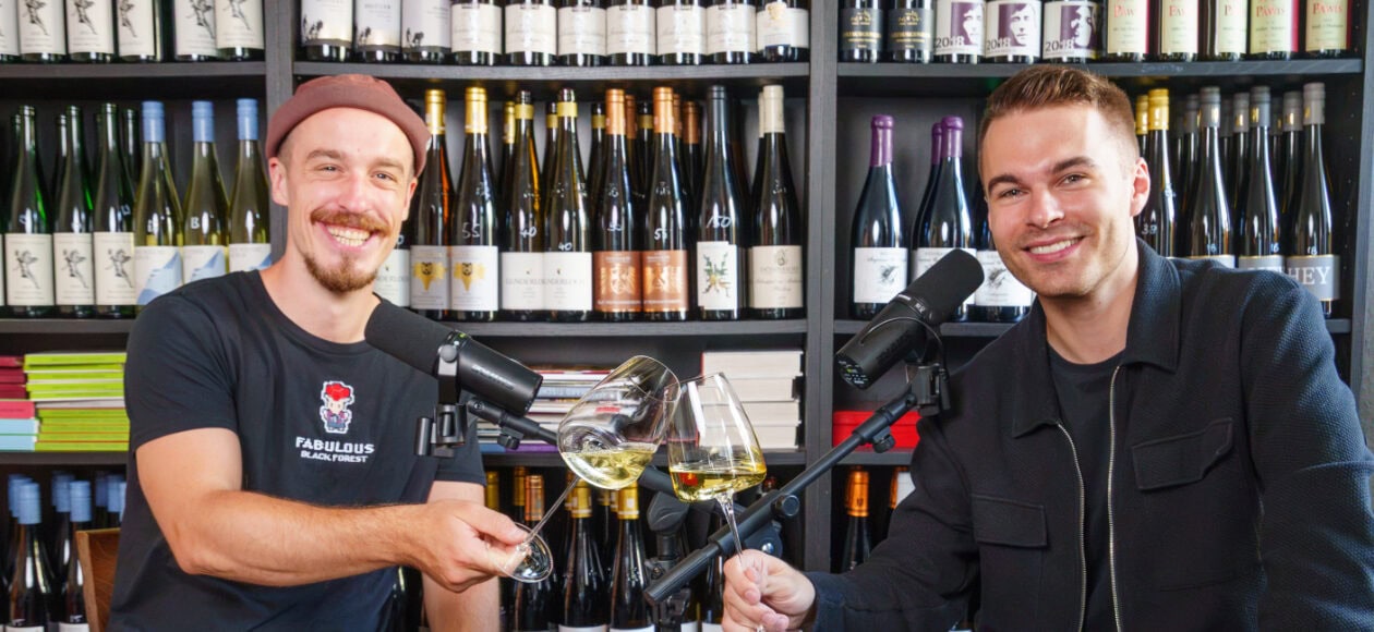 Zwei Männer sitzen vor einem Weinregal und stoßen mit einem Glas Weißwein an.