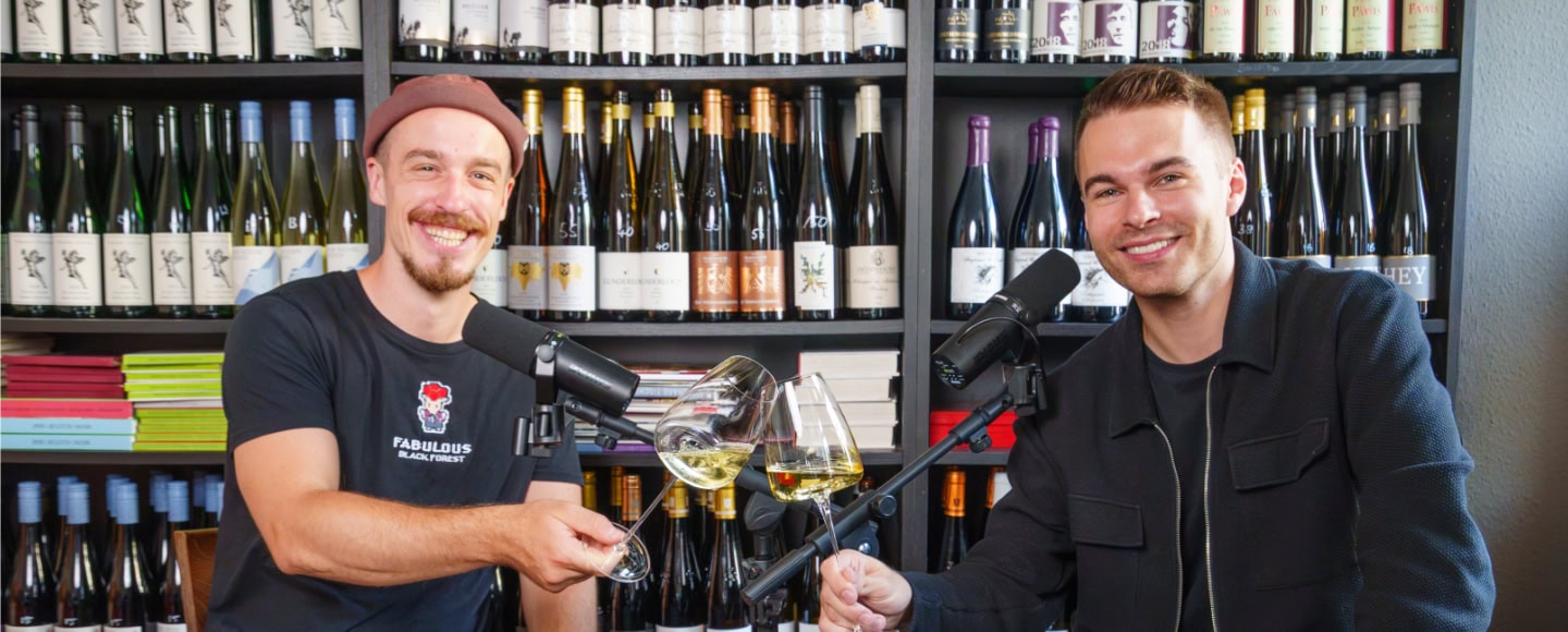Zwei Männer sitzen vor einem Weinregal und stoßen mit einem Glas Weißwein an.