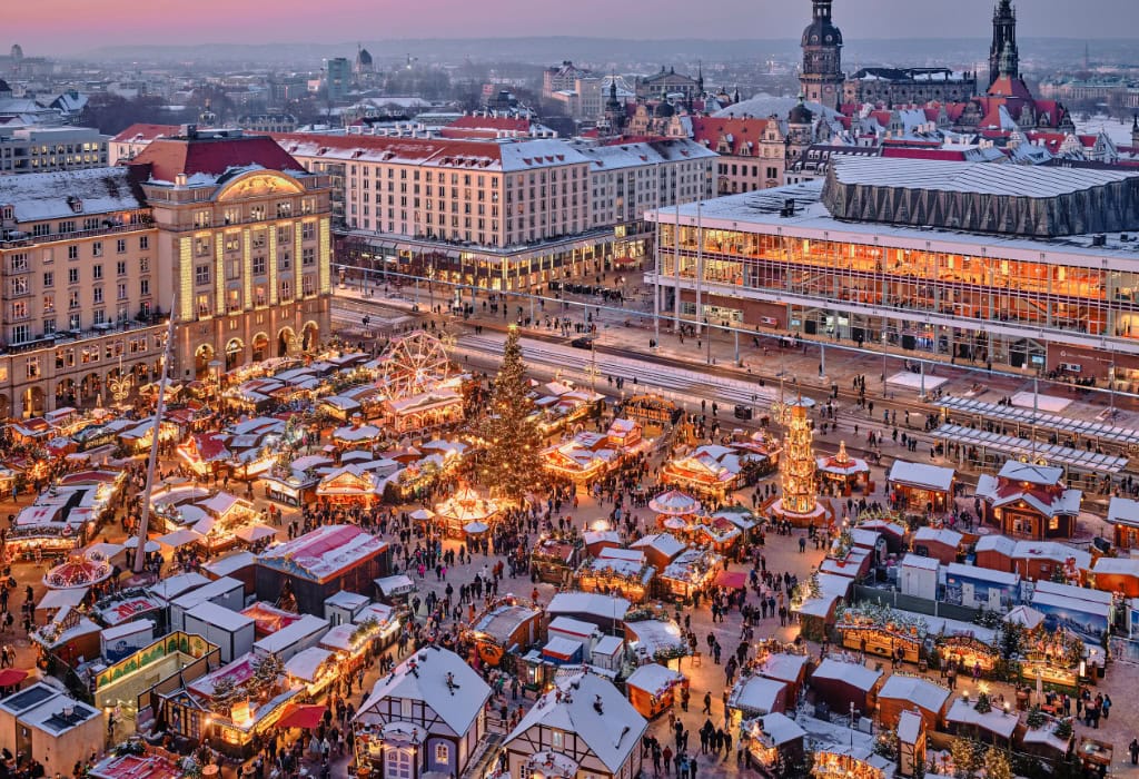 Blick von oben auf den verschneiten Dresdner Striezelmarkt mit festlich beleuchteten Marktständen, Weihnachtsbaum und historischer Altstadt im Hintergrund.